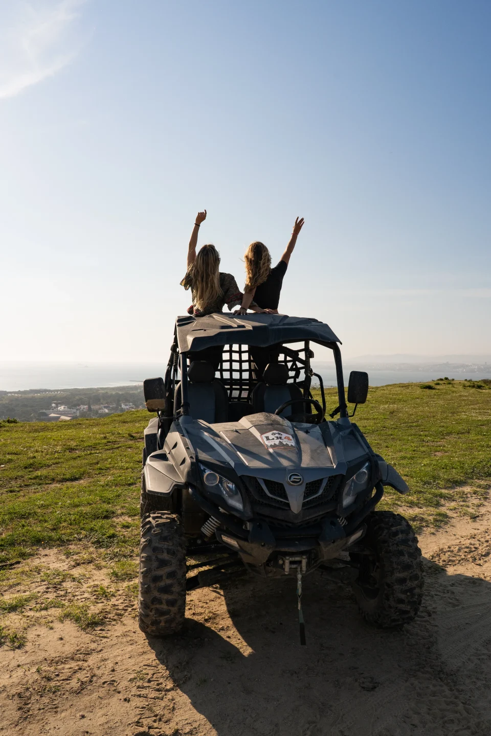 Two riders celebrating with arms raised on a buggy at a coastal hilltop